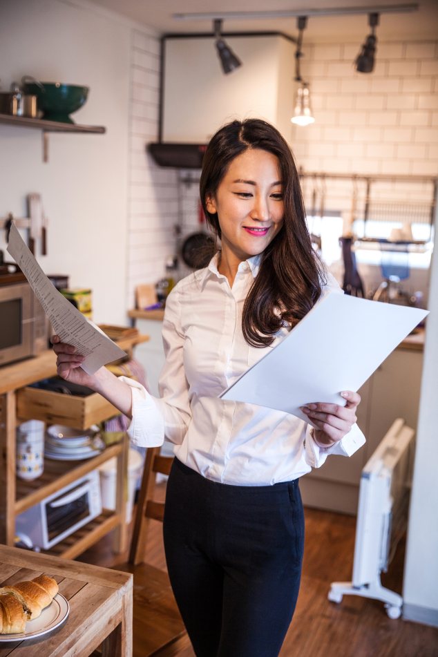 Woman in a white shirt and black pants reviewing papers in a kitchen with a microwave and pastries on a table.