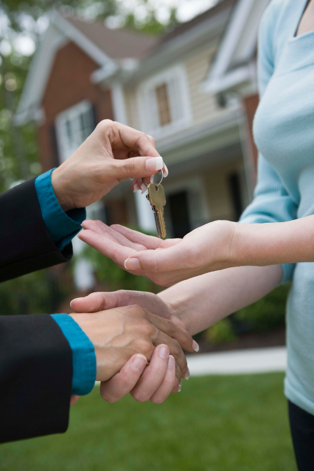 Person handing keys to another person, shaking hands in front of a house.