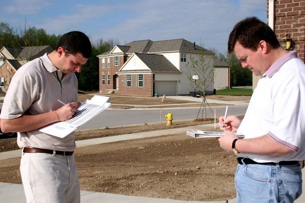 Two men writing on clipboards in front of a new house in a suburban neighborhood.