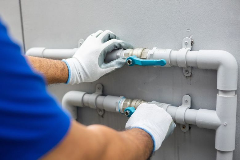 A person wearing gloves adjusts the blue handles of valves on gray industrial pipes mounted to a wall.