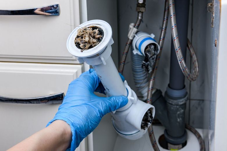 A gloved hand holds a dirty sink drain pipe, showcasing a severe blockage inside a kitchen cabinet.