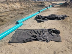 Blue PVC pipes lie across a sandy construction site with pieces of black geotextile fabric spread on the ground.