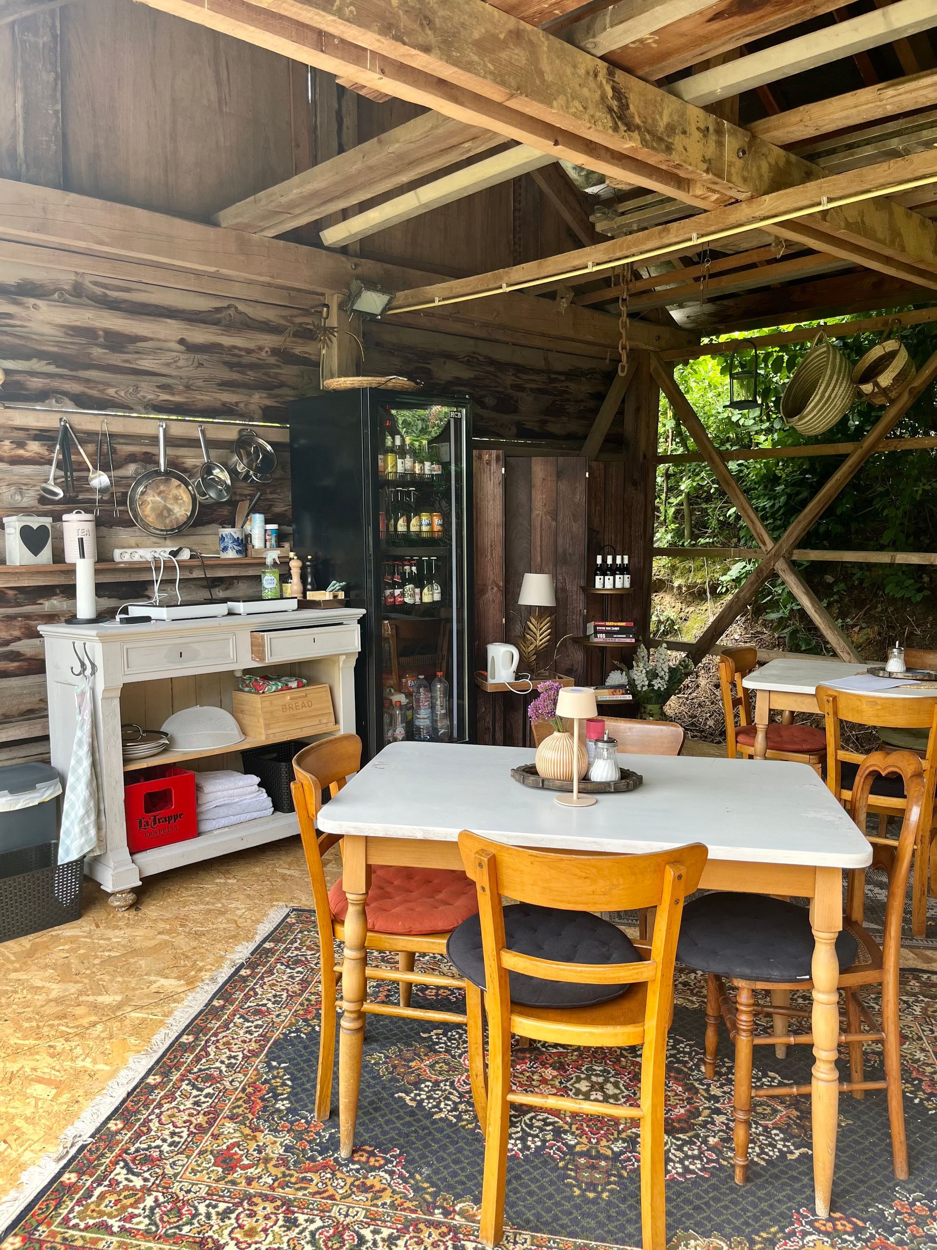Rustiek café met tafels, stoelen en vloerkleden. Een koelkast en schappen met producten zijn zichtbaar.