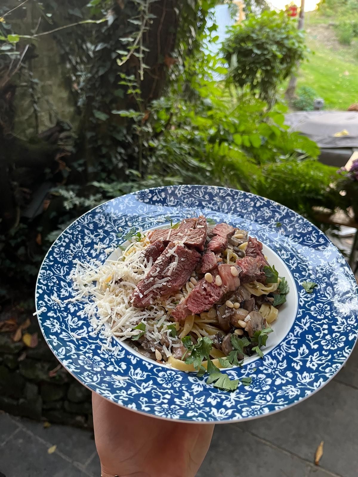 Een bord met biefstuk, pasta en kaas, buiten geserveerd te midden van groen gebladerte.