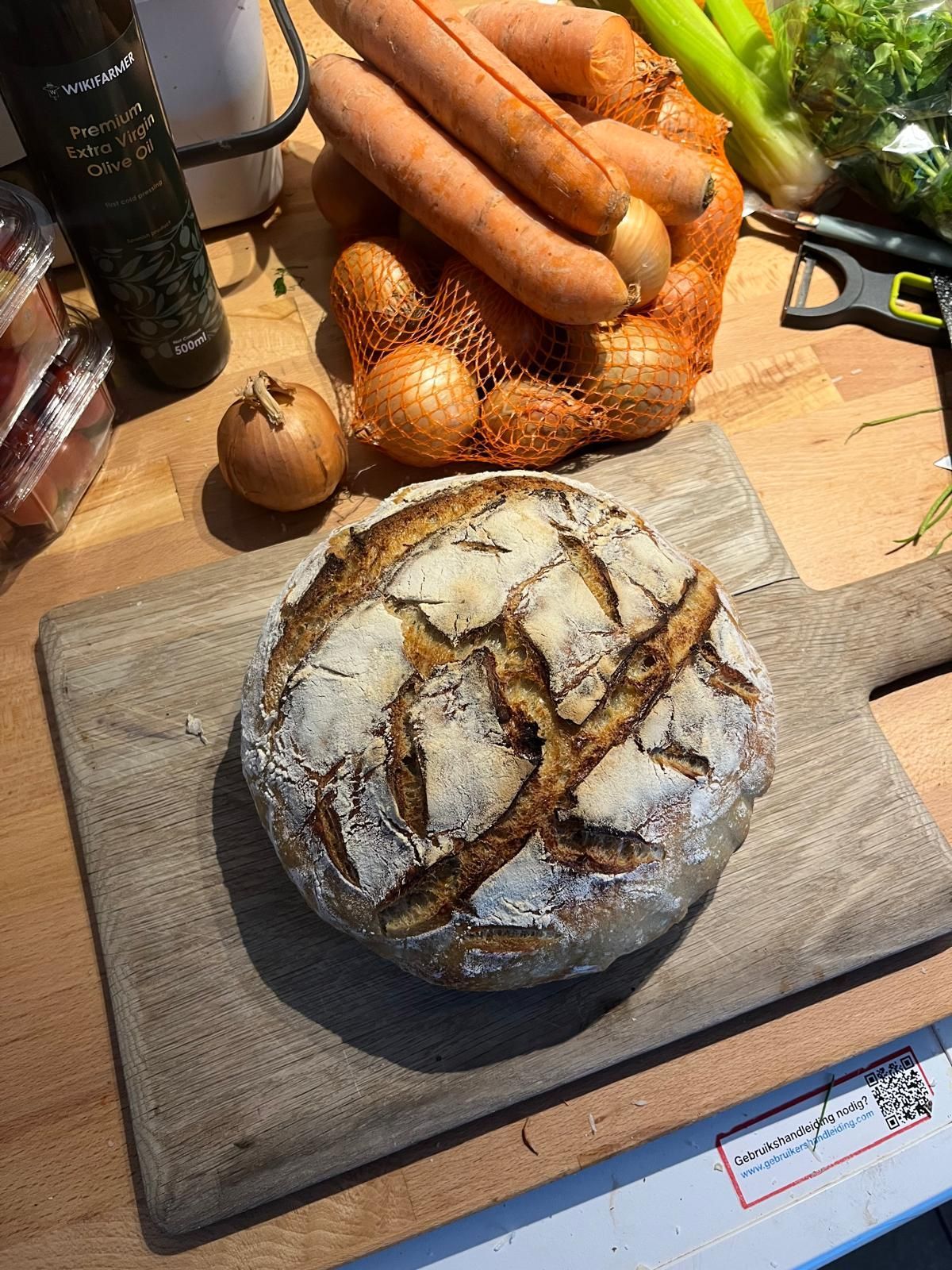 Rond brood op een houten plank, met groenten op de achtergrond: wortels en uien.