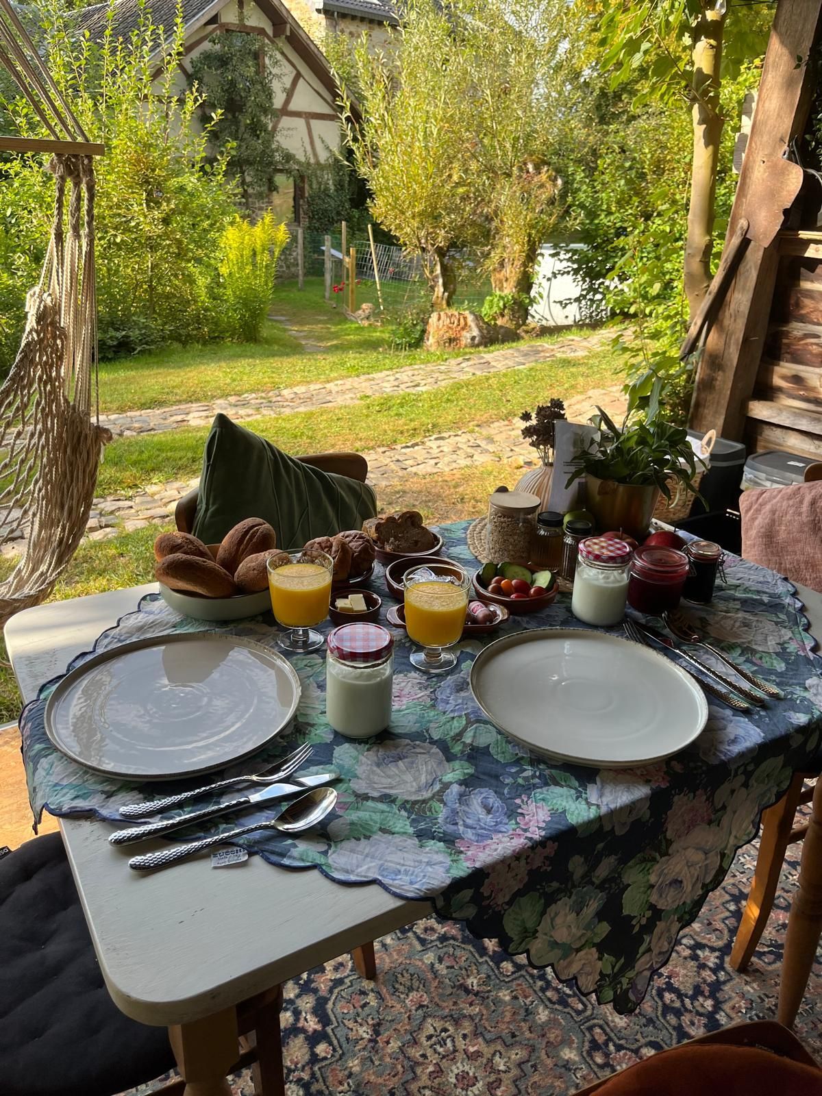 Ontbijttafel buiten, met croissants, sap, yoghurt en fruit. Op de achtergrond zijn een hangmat en een tuin te zien.