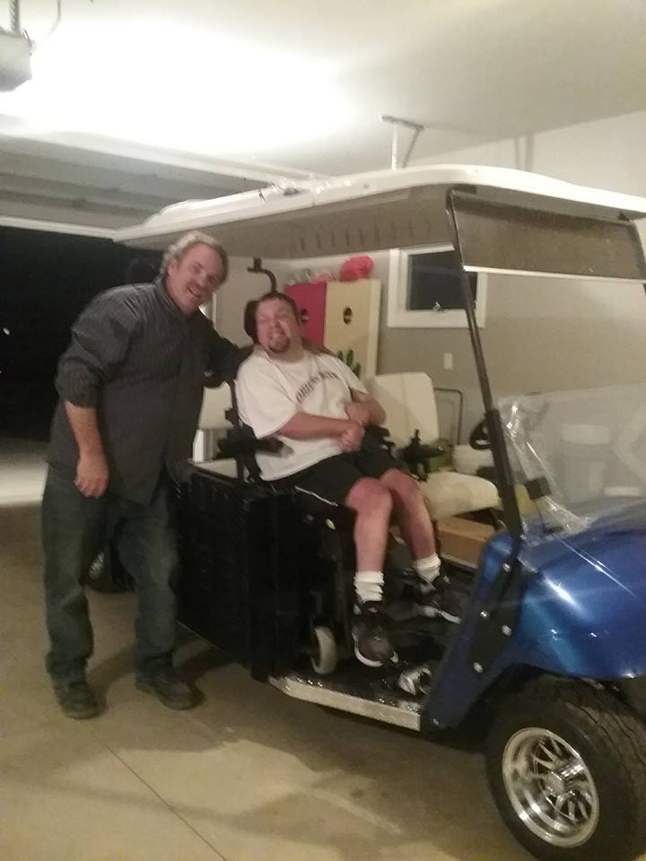 A man in a wheelchair is standing next to another man in a golf cart