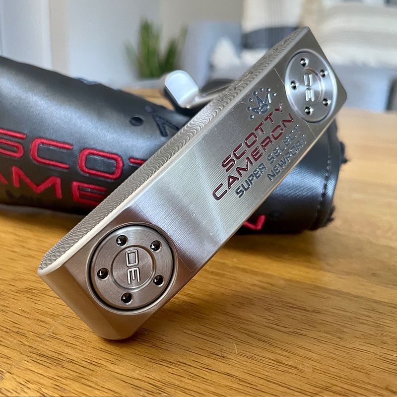 Silver Scotty Cameron Super Select Newport putter on a wooden surface, with a black headcover.