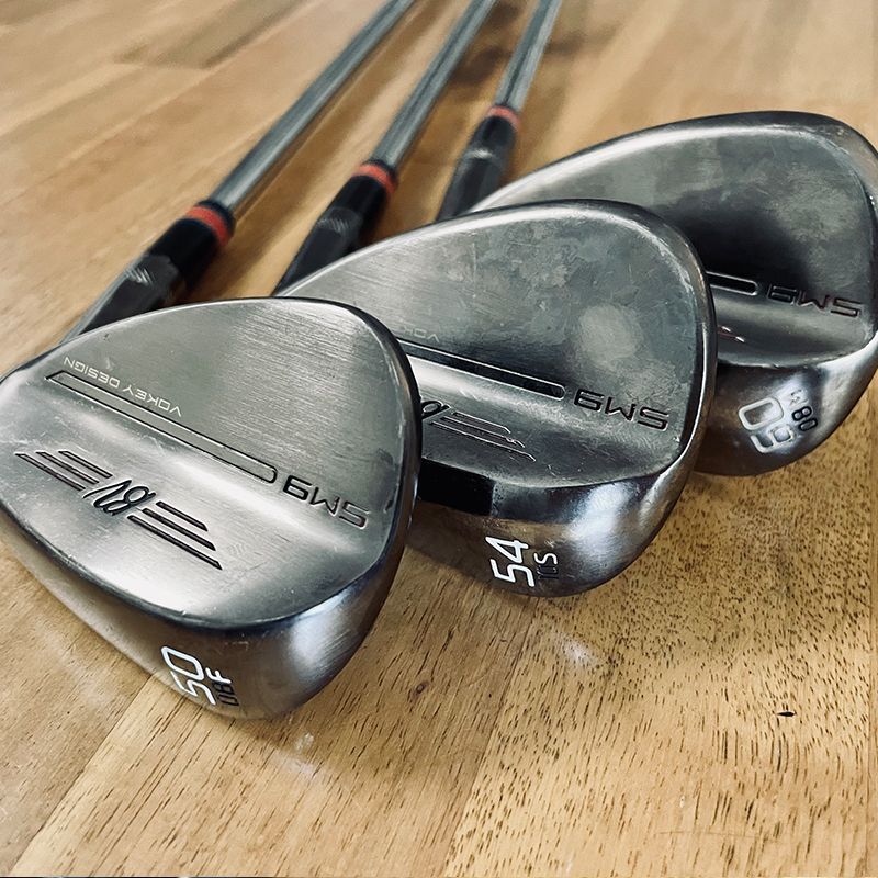 Four golf wedges with steel shafts and black grips, lying on a wooden surface.