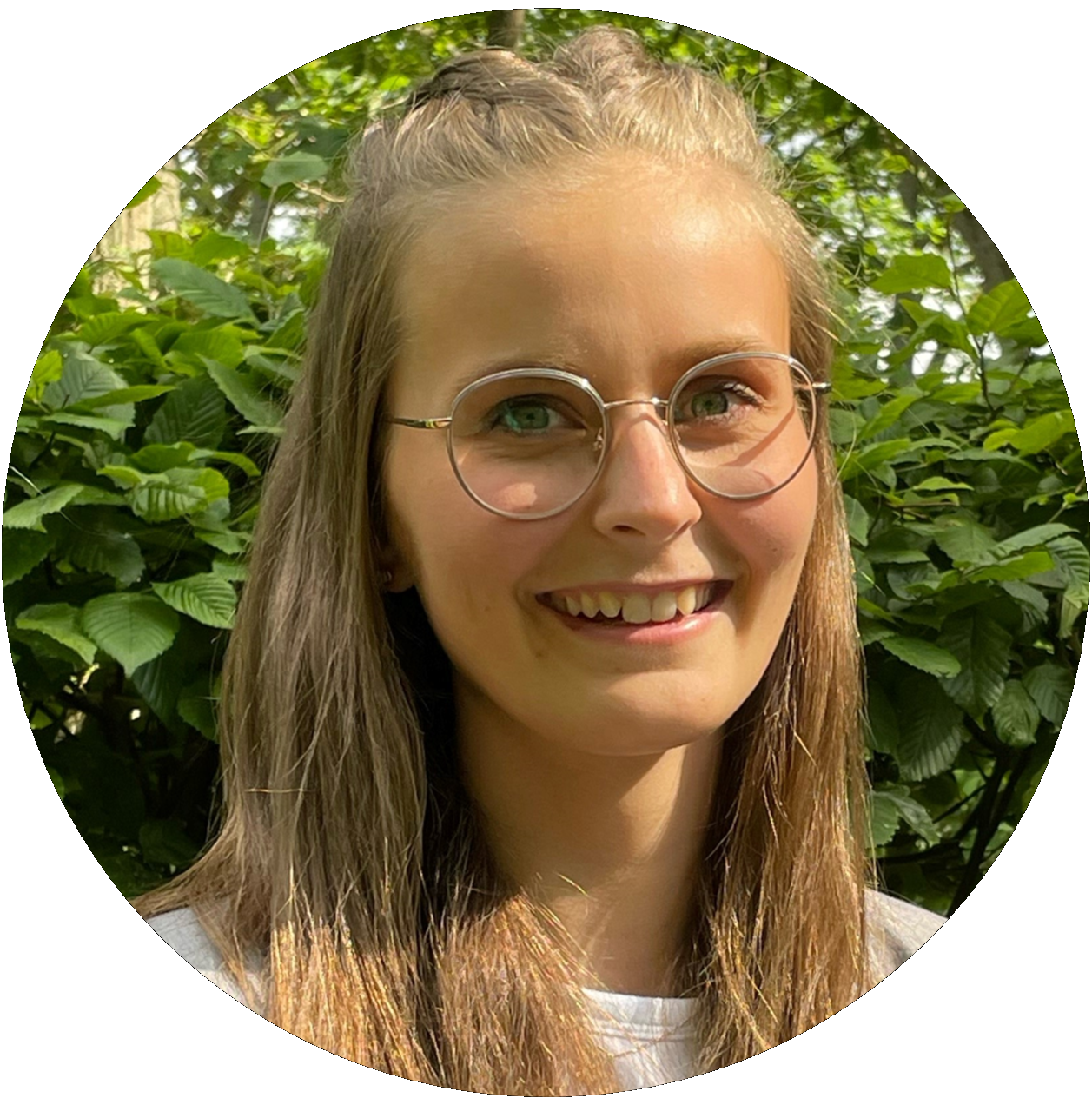 Junge Frau mit Brille, lächelnd, halb hochgestecktes Haar, im Freien vor Grün.