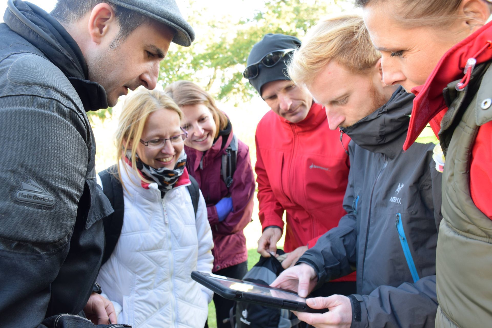 Gruppe von Menschen schaut im Freien auf ein Tablet. Einige lächeln, andere konzentrieren sich. Parklandschaft, bewölkter Himmel.
