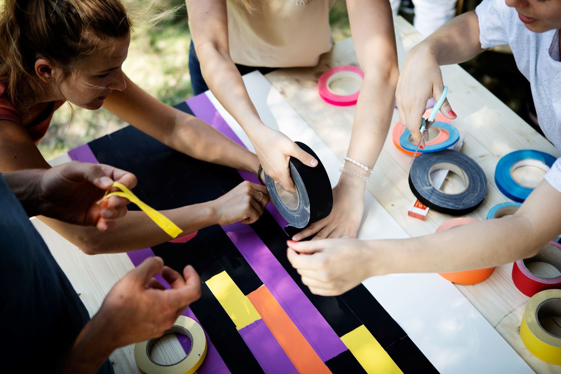 Menschen dekorieren draußen einen Holztisch mit buntem Klebeband; Hände arbeiten mit Klebebandrollen an dem Projekt.
