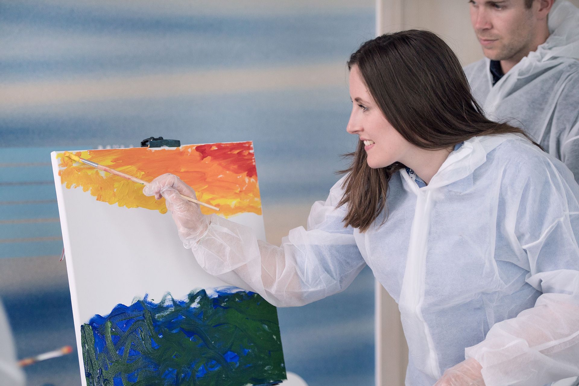 Frau malt mit einem Pinsel eine Landschaft. Mann schaut zu. Beide tragen Schutzanzüge.