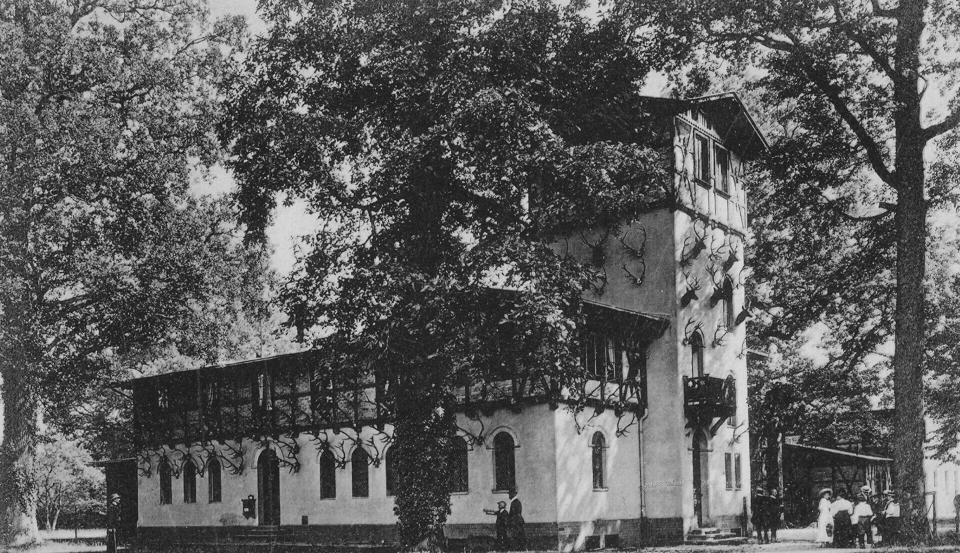 Ein weißes Gebäude mit einem Turm, umgeben von Bäumen. Auf dem Dach verläuft ein Balkon, und draußen haben sich Menschen versammelt.