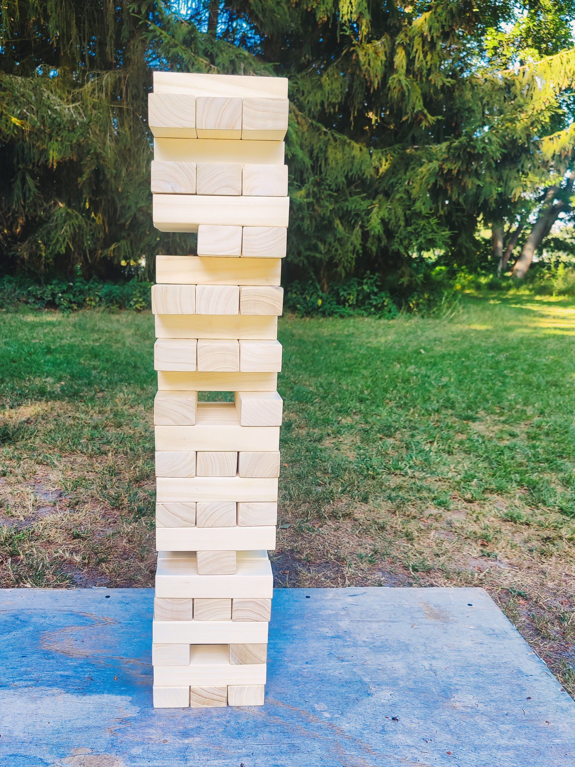 Ein riesiger Jenga-Turm aus hellen Blöcken steht auf einer Betonplatte in einer grasbewachsenen Außenumgebung.
