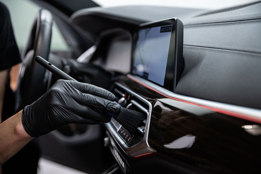 Person wearing black gloves cleaning a car's interior dashboard with a brush.
