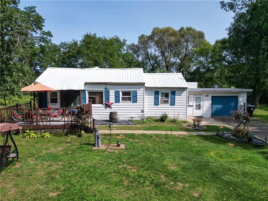 White house with a porch, blue shutters, and garage set in a grassy yard surrounded by trees.