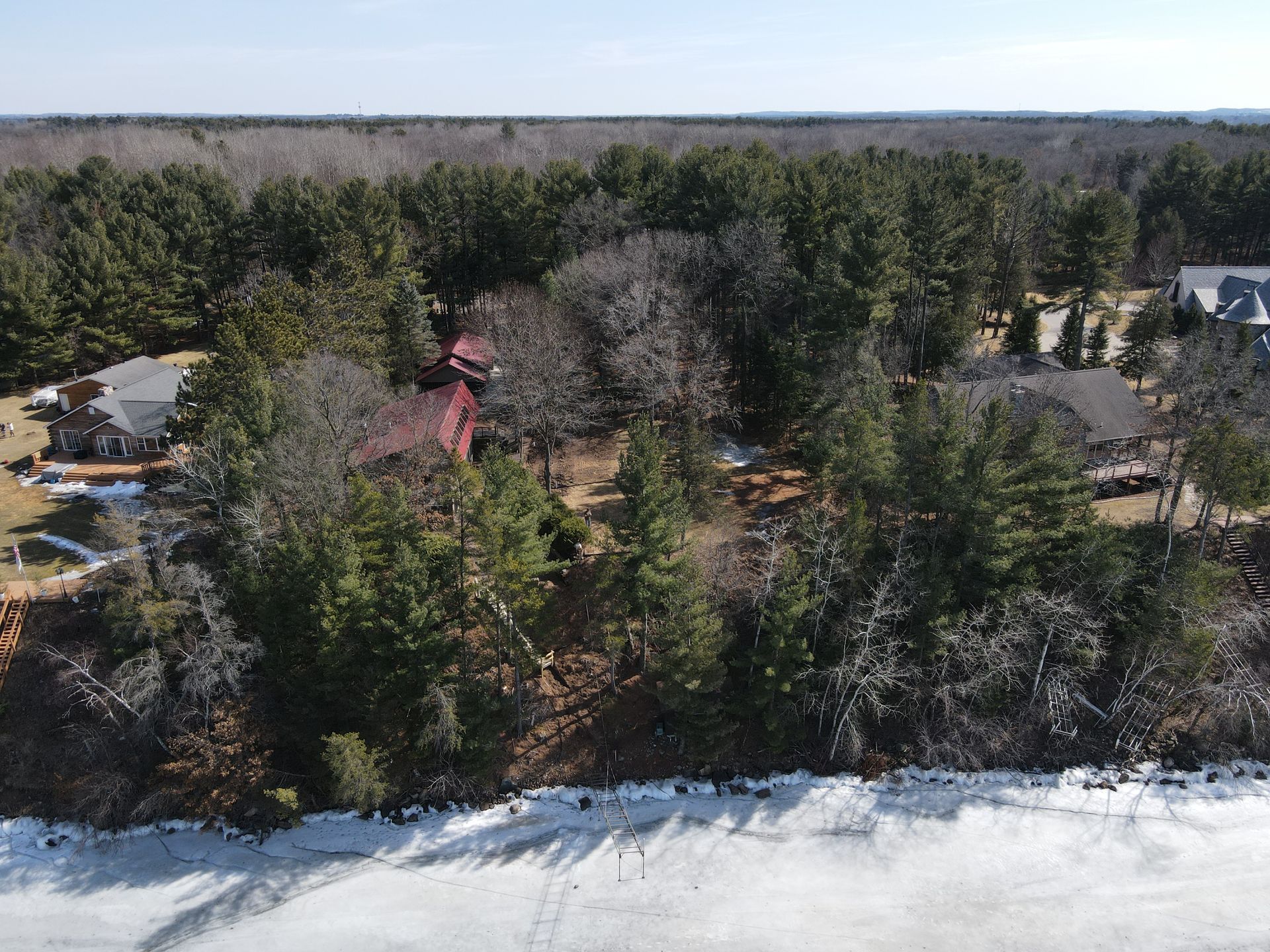 Aerial view of homes bordering a frozen lake, surrounded by evergreen and bare trees under a bright sky.