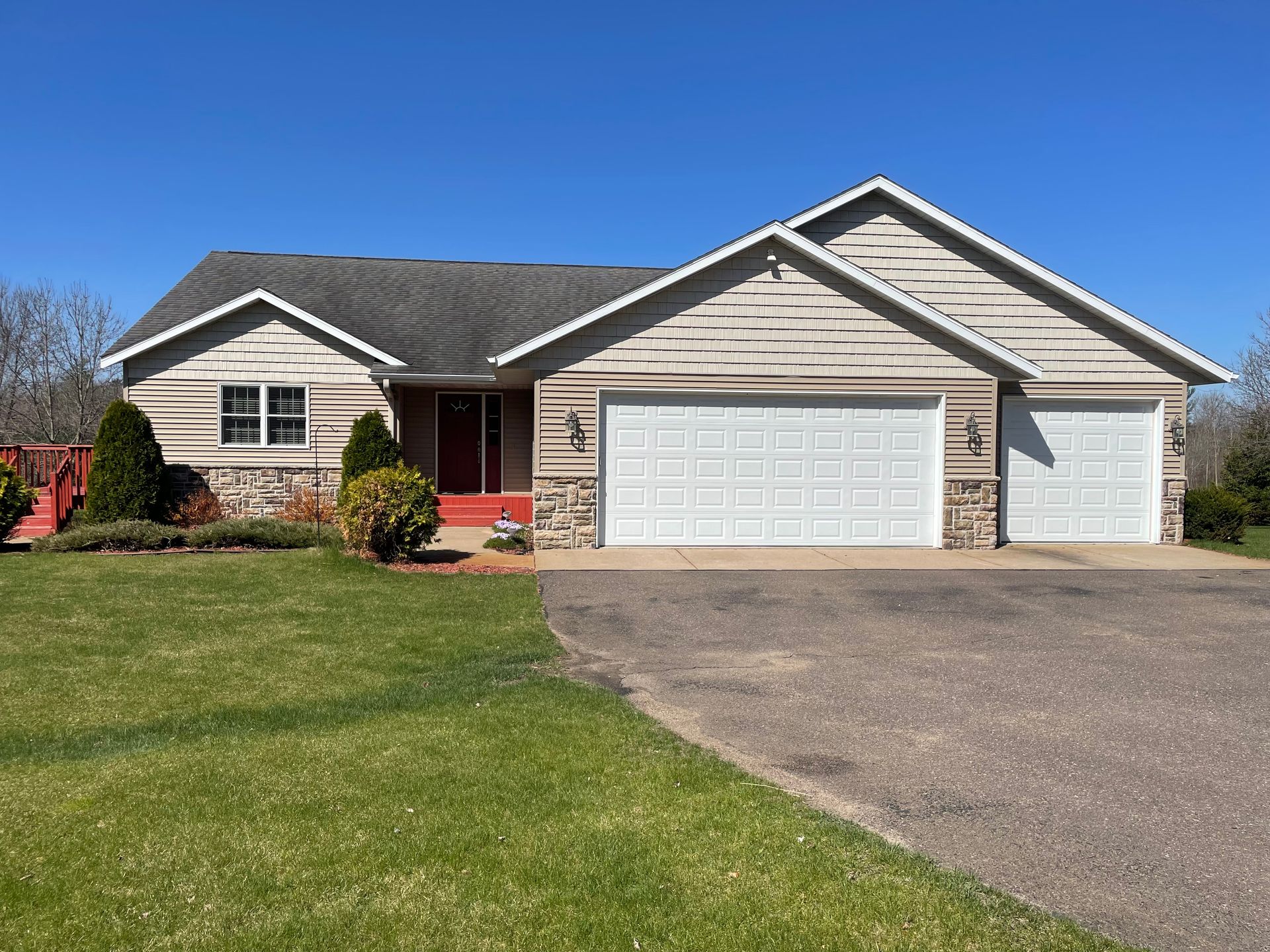 Ranch-style house with attached garage, beige siding, red door, and green lawn under a blue sky.