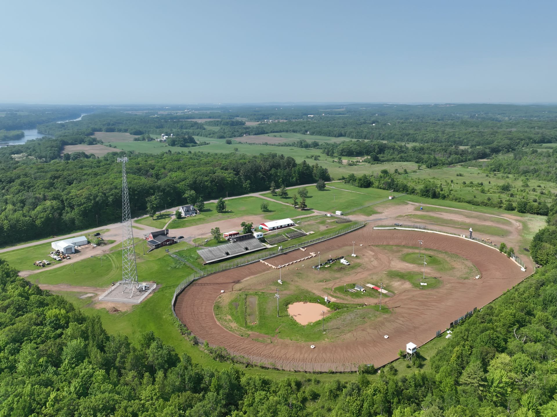 Aerial view of a dirt race track surrounded by trees and a green landscape under a blue sky.