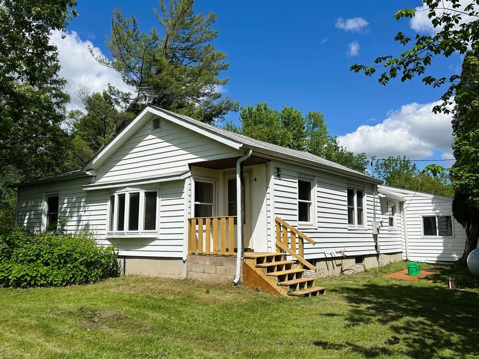 White house with wooden porch and steps, set in a grassy yard under a blue sky.