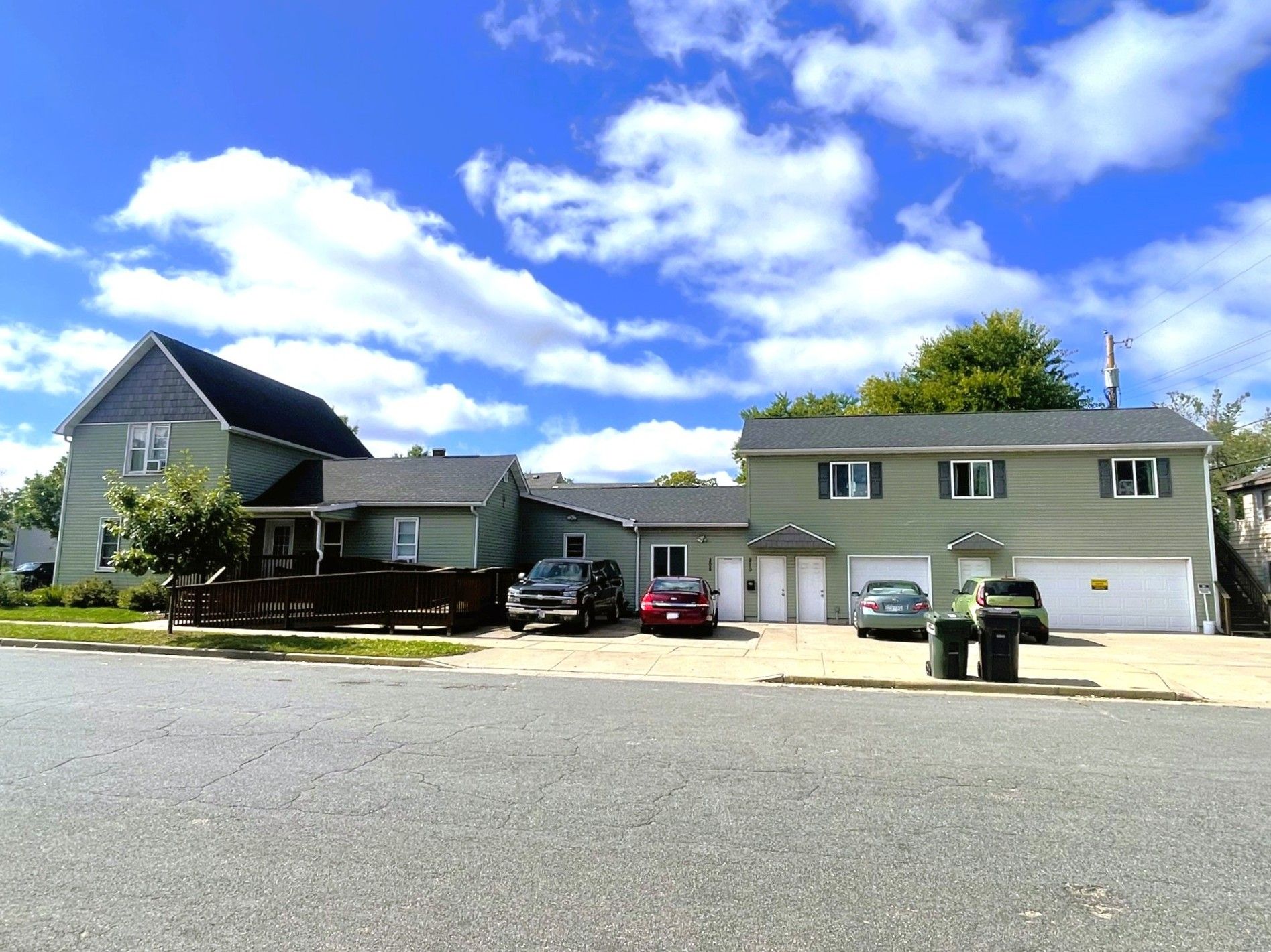 Multi-unit building with green siding, two-story garage, cars parked in front, blue sky with clouds.