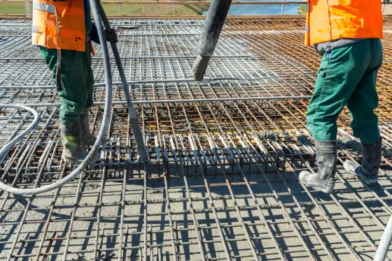 Construction workers pouring concrete onto a rebar grid, wearing orange vests and green pants.