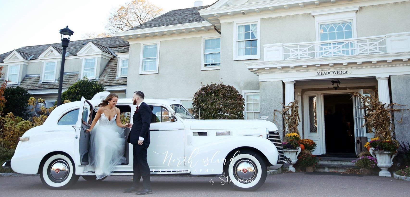 A bride and groom are standing next to a white limousine in front of a large white house.