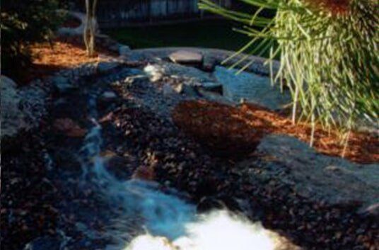 Water cascading down a rocky stream in a landscaped garden, with lush greenery.