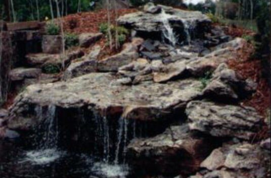 Waterfall cascading over layered rocks, surrounded by mulch and trees.