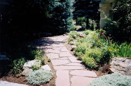 Stone path winds through a garden with lush plants and trees on either side.