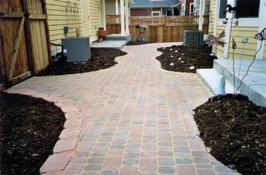 Brick pathway winding through mulch beds, flanked by wooden fences and buildings.