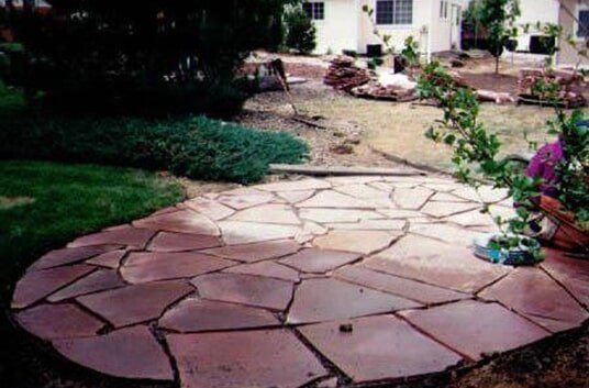 Flagstone patio in a yard, surrounded by grass, plants, and a house in the background.