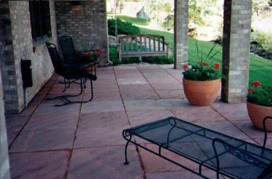Brick patio with wrought iron furniture and planters with red flowers. A wooden swing is in the background.