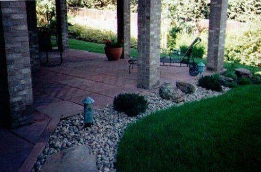 Patio with brick columns, cobblestone ground, and garden bed with grass and rocks.