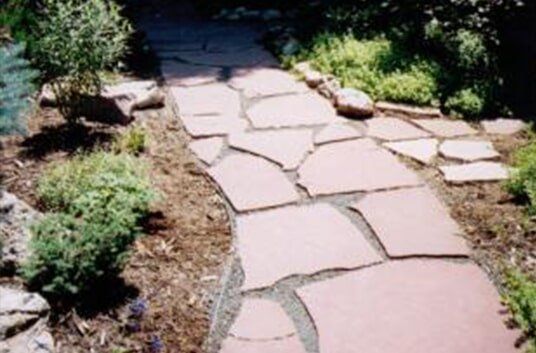 Stone pathway through a garden, with reddish-brown stones and green plants on either side.