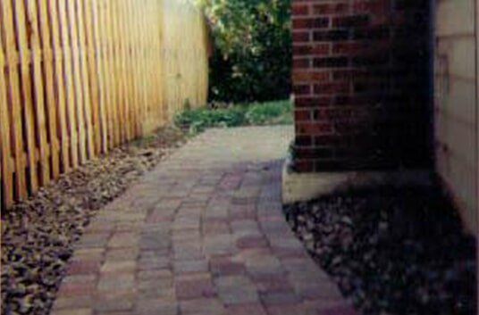 Brick walkway between a wooden fence and a brick wall. Gravel borders each side.