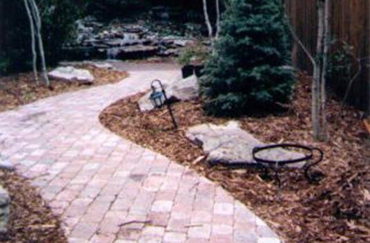 Brick pathway winding through a landscaped yard with trees, mulch, and a fire pit.