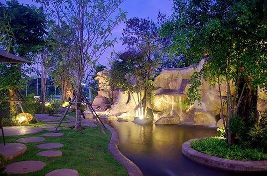 Scenic outdoor garden with a pond, rock formations, trees, and illuminated pathways.