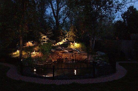 Night view of a illuminated pond and waterfall surrounded by trees and a brick pathway.