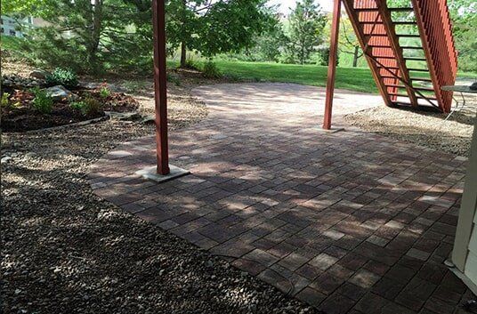 Brick patio with stairs and brown support beams in a yard with trees.