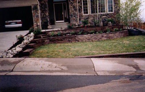 House with stone facade, landscaped front yard, concrete steps, garage on the left, and grass and flowers in the yard.