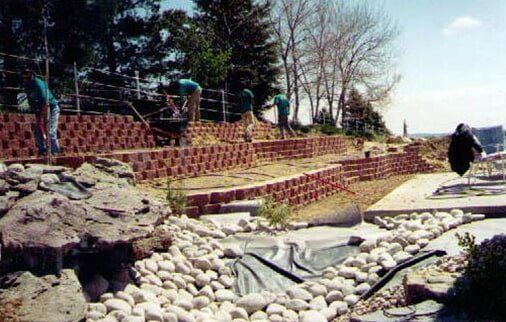 Workers building terraced retaining walls; a rocky stream bed in the foreground.