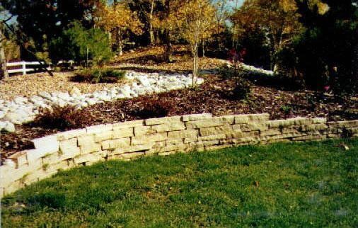 A stone retaining wall curves along a grassy lawn, with a wooded hillside in the background.