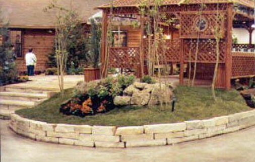 A wooden gazebo and rock garden in a yard with a stone retaining wall, a person in the background.