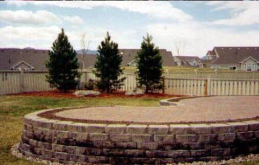 Brick patio with curved retaining wall, trees, fence, and houses in the background.