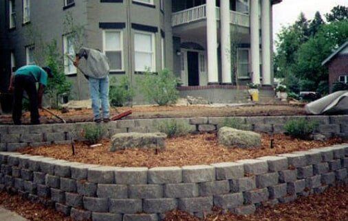 Men working on a landscaped yard with a stone retaining wall in front of a house.