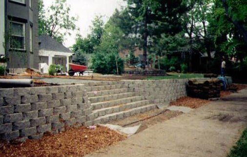 Stone retaining wall with steps leading up, next to a sidewalk and house.