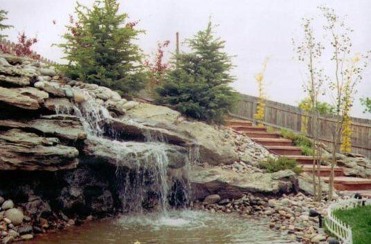 Waterfall cascading over rocky ledge into small pond, beside stairs and fence.