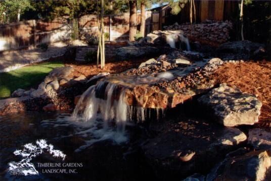 Water cascades over rocky falls into a pond; brown mulch, green grass, and trees.